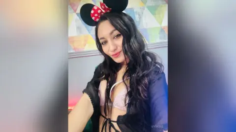 Smiling woman with long dark hair wearing Minnie Mouse ears and a black lace outfit in a bright room.