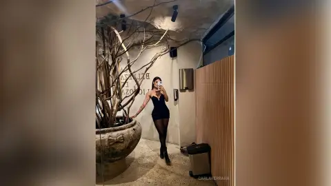 Elegant woman in a black cocktail dress and tights posing in a modern, minimalist interior with natural decor.