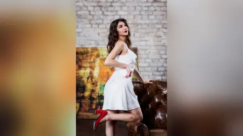 Elegant woman in a white cocktail dress and red heels posing confidently against a brick wall background.