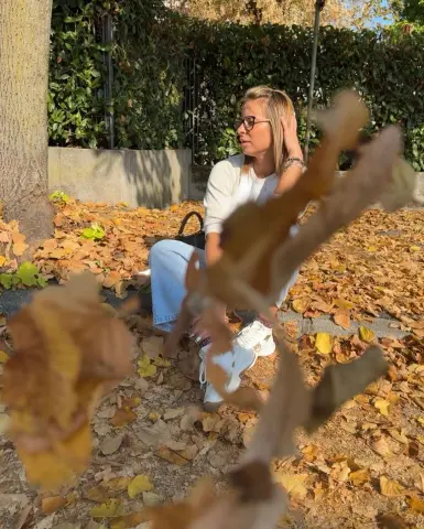 Woman in white sweater and jeans sitting in autumn leaves outdoors on a sunny fall day.