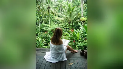 MilaSwan — MilaSwan sits on a wooden deck in a white off-the-shoulder shirt while reading a book overlooking a lush tropical jungle.