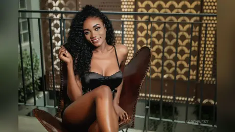 Smiling woman in a black cocktail dress sitting on a leather chair on a balcony with decorative railings.
