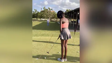 Young woman in a pink tank top and grey skirt playing golf on a sunny green course.