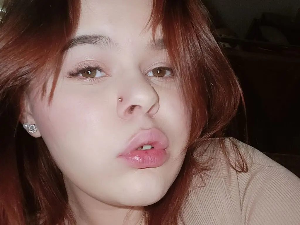 Close-up portrait of a young woman with reddish-brown hair and nose piercing in soft indoor lighting.