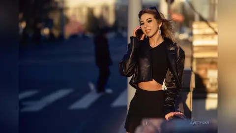 Smiling woman in a black leather jacket and crop top posing outdoors during a warm golden hour sunset.