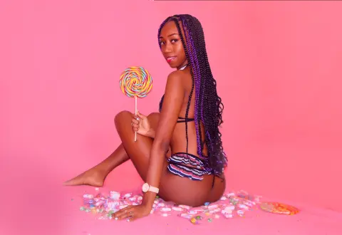 Smiling Black woman with long braids in a striped bikini holding a colorful lollipop against a pink background.