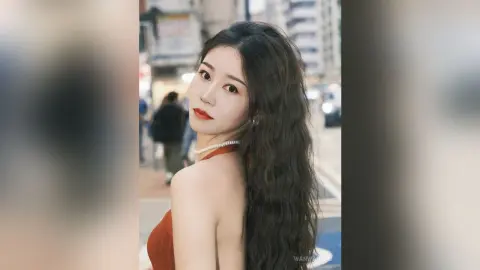 Young woman with long wavy hair wearing a red dress and pearl necklace in a bright urban street setting.