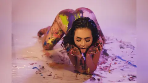 Artistic woman covered in colorful paint lying on a white floor in a dreamy, creative studio setting.