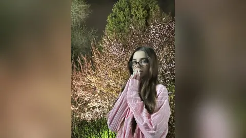 Young woman in a pink striped shirt posing thoughtfully in a garden with blooming trees at night.