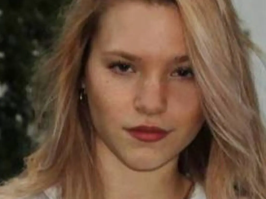 Blonde woman with freckles wearing a white top in a soft, natural outdoor setting.
