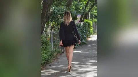 Blonde woman in a black mini dress and heels walking down a sunny tree-lined path.