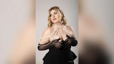 Elegant woman in a black dress with pearl body jewelry and sheer gloves posing in a studio setting.