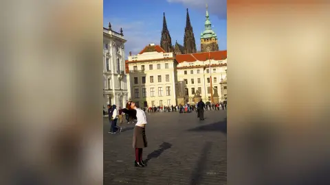 Young woman in a white shirt and grey skirt standing in a sunny historic European plaza near a cathedral.