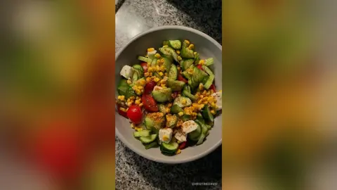 ChristineDavis prepared a fresh salad with sliced cucumbers, cherry tomatoes, corn, and cubes of white cheese in a grey bowl.