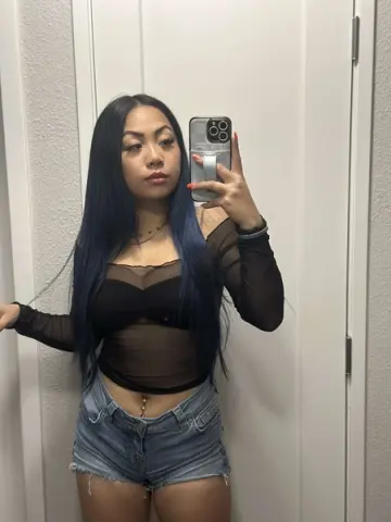 Young woman with long blue hair in a black sheer top and denim shorts taking a mirror selfie at home.