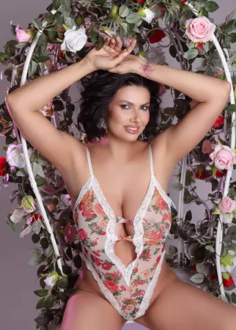 Smiling woman in floral lace bodysuit sitting in a romantic rose garden swing setting.