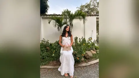 Young woman in a white summer maxi dress holding a beige handbag in a lush tropical garden setting.