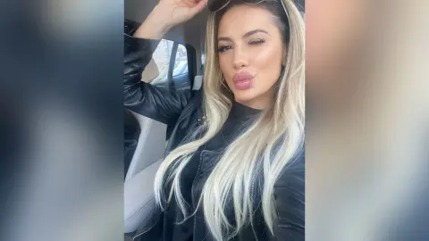 Glamorous blonde woman in a black leather jacket posing inside a car with a confident expression.