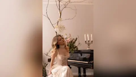 Elegant woman in a white lace corset gown sitting by a piano in a dreamy, romantic indoor setting.