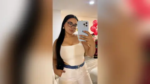 NatalyBloom poses in a white tank top and white pants with a blue belt, wearing glasses and holding a phone for a mirror selfie.
