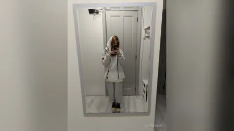 Young woman in a cozy white hoodie and grey sweatpants taking a casual mirror selfie in a bright room.