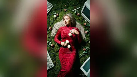Elegant woman in a red velvet gown lying on green moss surrounded by white roses and vintage technology.