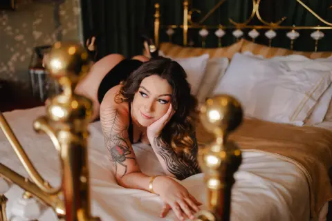 spicyspiders21 lies on her stomach on a bed, showing her tattooed arms and chest while resting her chin on her hand.