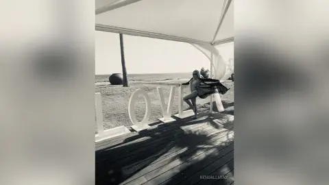 KendallMinx poses in a black bikini on a wooden deck next to large white LOVE letters under a white canopy.
