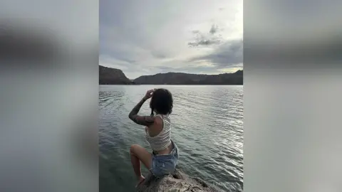 Woman in a striped tank top and denim shorts sitting on a rock overlooking a vast lake and mountains.
