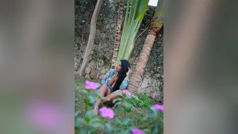 MissyJolie poses on a grassy lawn wearing a patterned blue lace bodysuit, surrounded by pink flowers and palm trees.