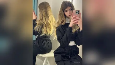 Young woman with long wavy blonde hair wearing a black jumpsuit taking a mirror selfie in a bright room.