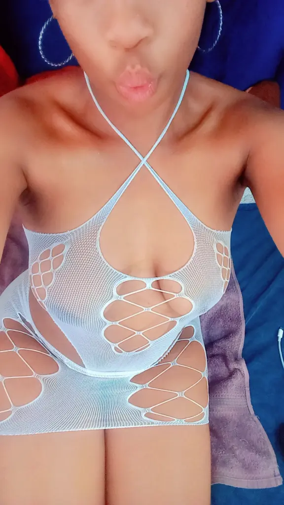 MakeMeWantIt93 sits on a white tufted sofa wearing a blue tank top and dark shorts, showing off her long dark hair and tanned skin.