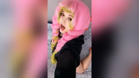 EstherTrix poses on a bed with her tongue out, wearing a black top and bright pink hair with green highlights.