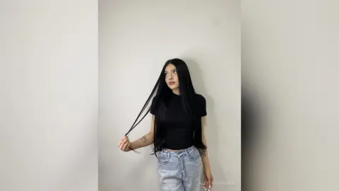 AlettaFlorence poses against a white wall, wearing a black t-shirt and light blue jeans while holding a long strand of her long black hair.