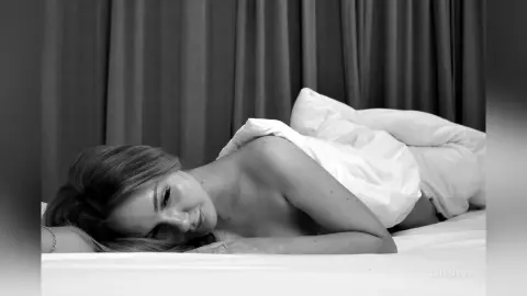 Relaxed woman lying in bed under white linens in a moody black and white indoor setting.