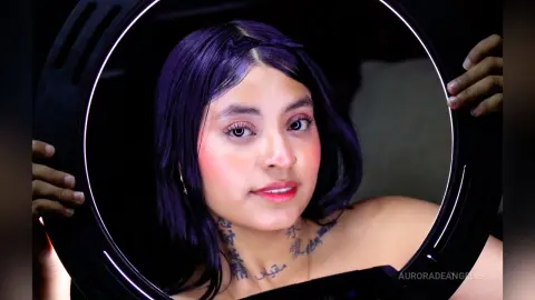 Young woman with purple hair and neck tattoos looking into a ring light in a moody, dark setting.