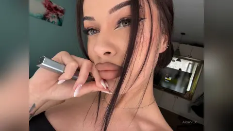 AriaVex poses close-up, holding a silver vape pen near her lips with long white manicured nails and dark hair framing her face.