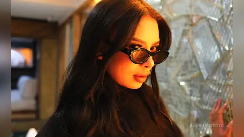 Stylish woman with long dark hair wearing black sunglasses in a warm, moody indoor setting.