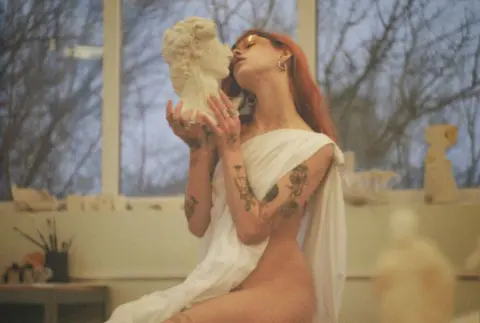 AlisaMoss is a redheaded woman with tattoos, wearing a white cloth draped over her body while posing with a white bust in a studio.