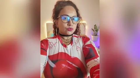 Woman with curly hair and glasses wearing a red and white patterned top in a cozy bedroom setting.