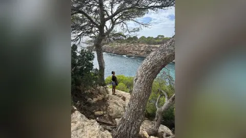 LucyMartin stands on a rocky cliffside overlooking a bright blue ocean, surrounded by large pine trees.