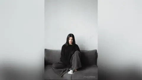 Young woman in a black sweater and grey trousers sitting on a grey sofa in a minimalist white room.