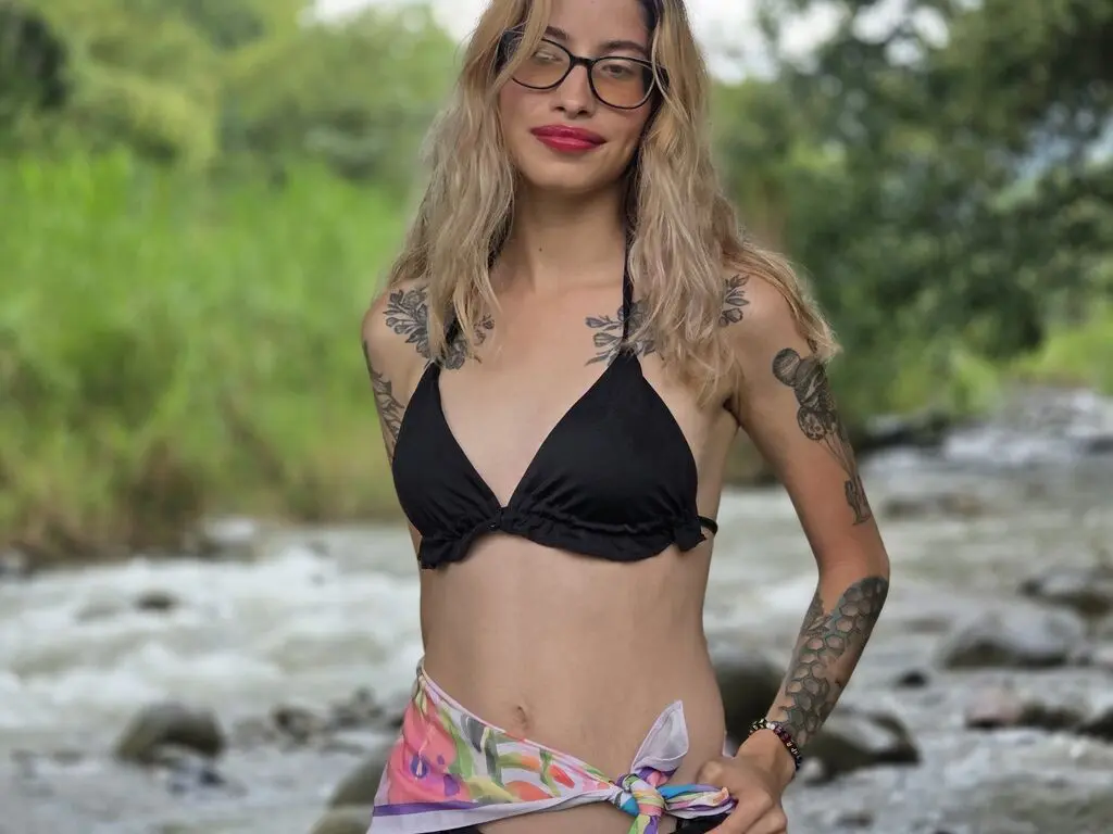 Smiling blonde woman in a black bikini top standing by a flowing river in a lush green forest setting.
