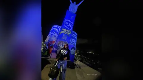 Woman in a black graphic sweatshirt standing before a colorful street art monument at night.