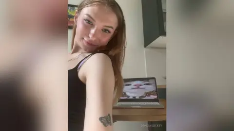 DahliaSackey smiles while wearing a black tank top, showing her long brown hair and a butterfly tattoo on her arm next to a laptop.
