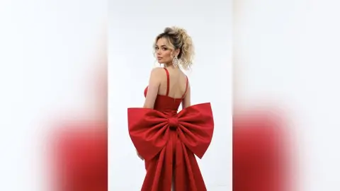 Elegant woman in a glamorous red evening gown with a large statement bow against a white background.