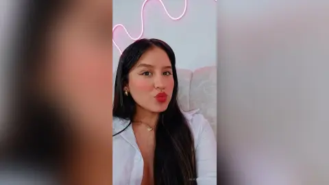 Young woman with long dark hair wearing a white shirt and red lipstick in a cozy room with neon lighting.