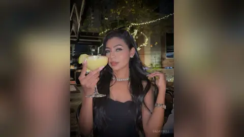Glamorous woman in a black dress holding a cocktail at an outdoor night lounge with fairy lights.
