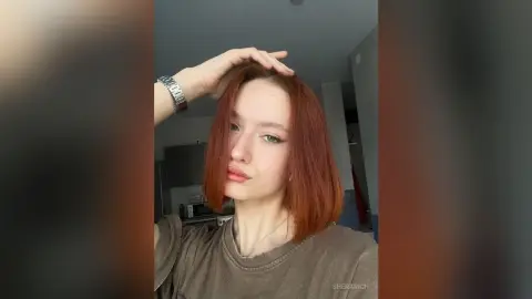 SheraRich poses with her red hair, wearing a brown t-shirt and a silver watch, looking directly at the camera.