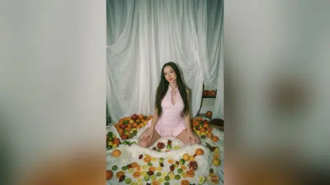 Young woman in a pink halter dress sitting amidst fresh fruits on white faux fur in a dreamy studio setting.